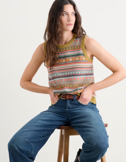 Woman wearing a patterned sleeveless top and blue jeans sitting on a stool against a white background