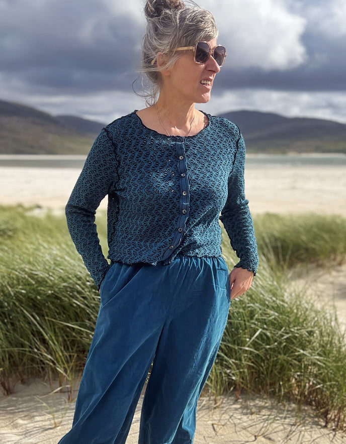 A woman wearing a dark teal patterned long-sleeve button-front top and matching teal pants, standing outdoors in a grassy area with mountains and a cloudy sky in the background.
