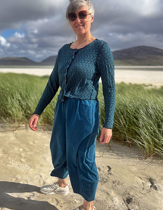 A woman wearing a teal patterned long-sleeve top and matching teal wide-leg pants stands on a sandy beach with grass and distant hills in the background.
