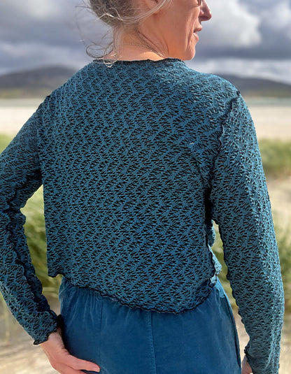 A woman wearing a textured teal blue long-sleeve top and matching blue skirt, photographed outdoors with a blurred natural landscape background.