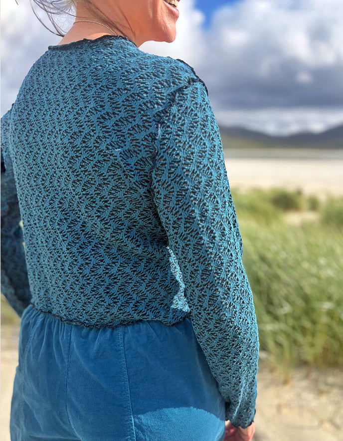 A woman wearing a teal blue textured knit long-sleeve top and matching blue skirt, photographed outdoors from behind against a blurred beach and sky background.