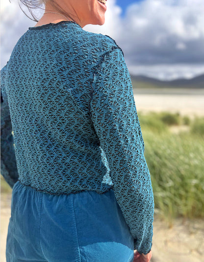 A woman wearing a teal blue textured knit long-sleeve top and matching blue skirt, photographed outdoors from behind against a blurred beach and sky background.