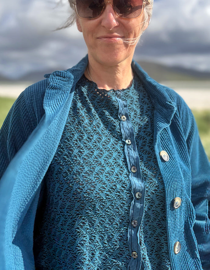 A close-up lifestyle shot of a woman wearing a textured blue button-up shirt and a matching blue corduroy cardigan, with a blurred outdoor landscape in the background.