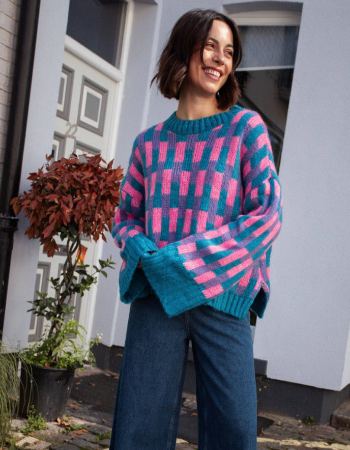 A woman wearing a colorful knit sweater with a pink and teal geometric pattern, paired with dark blue jeans, standing outdoors in front of a white house.
