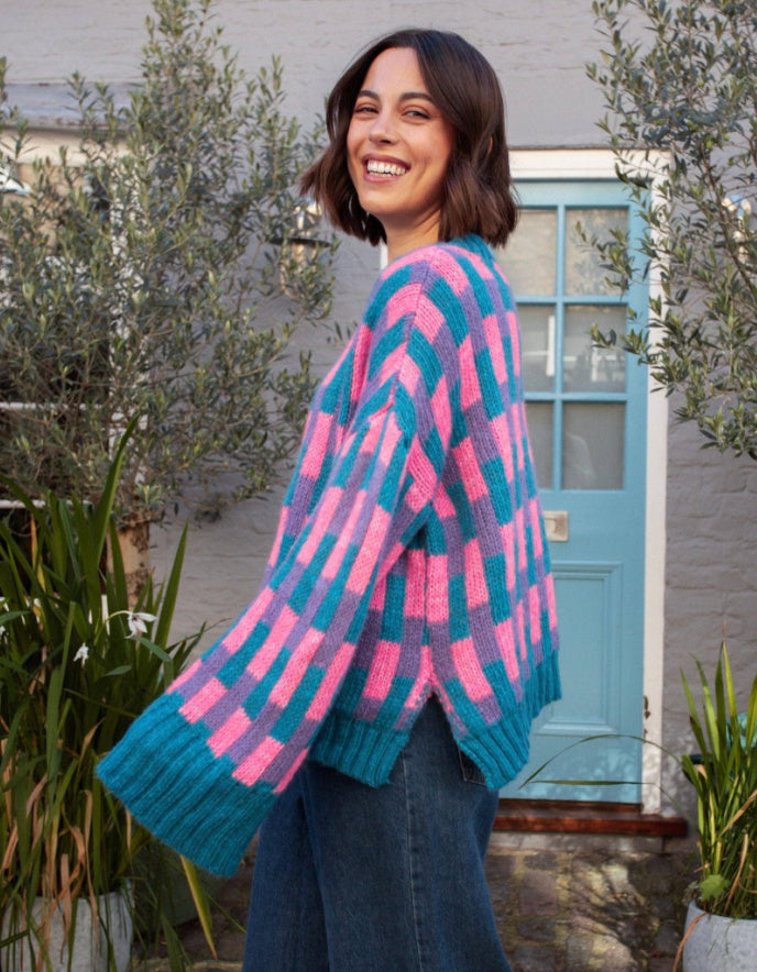 A woman wearing a chunky knit cardigan with a pink and teal chevron pattern, standing outdoors in front of a light blue door and potted plants.