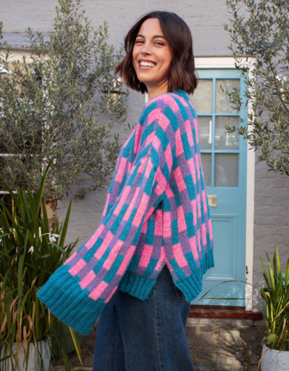 A woman wearing a chunky knit cardigan with a pink and teal chevron pattern, standing outdoors in front of a light blue door and potted plants.