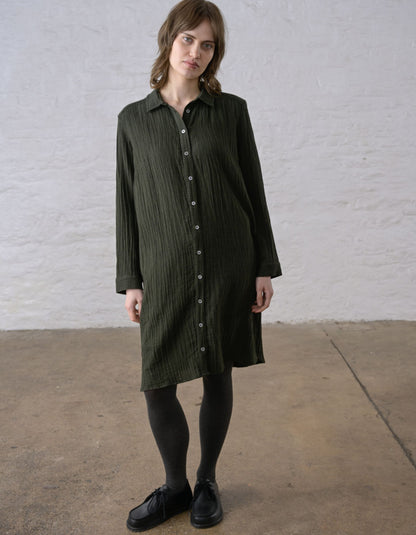 A woman wearing a dark olive green crinkled shirt dress with a button-down front, paired with black tights and black leather shoes, standing against a white brick wall.
