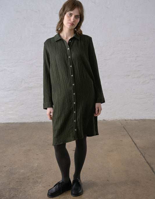 A woman wearing a dark olive green crinkled shirt dress with a button-down front, paired with black tights and black leather shoes, standing against a white brick wall.