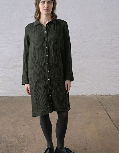 A woman wearing a dark olive green textured long-sleeve button-down shirt dress, paired with black tights and black shoes, standing against a white brick wall.