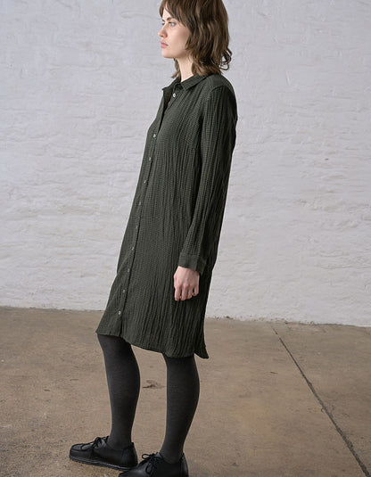 A woman wearing a dark olive green textured long-sleeve button-up shirt dress, paired with black tights and black shoes, standing against a white brick wall.