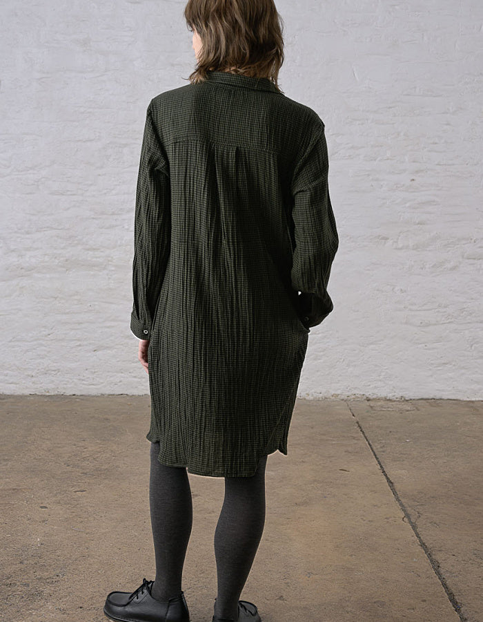 A person shown from the back wearing a dark olive green textured shirt dress, black tights, and black shoes, standing against a white brick wall and concrete floor.