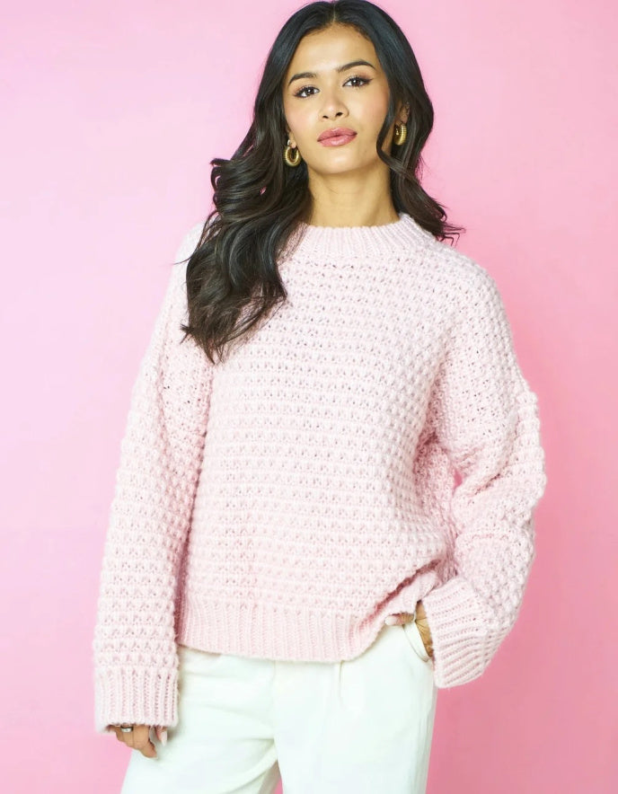 A woman wearing a light pink textured knit sweater and white pants, standing against a solid pink background.