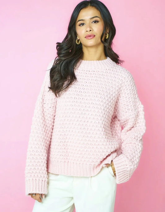 A woman wearing a light pink textured knit sweater and white pants, standing against a solid pink background.