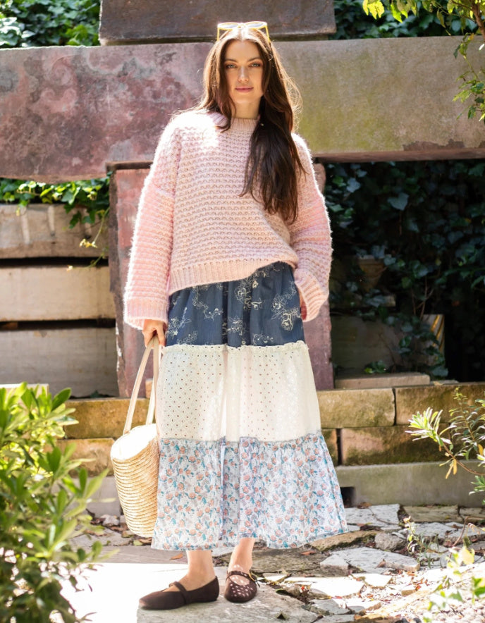 A woman stands outdoors in a garden setting wearing a chunky light pink knit sweater and a long patchwork maxi skirt with blue, white, and floral sections. She is holding a woven straw tote bag.