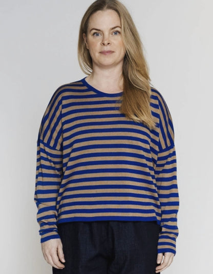 A woman wearing a blue and tan striped long-sleeve knit top, paired with dark pants, standing against a light gray studio background.