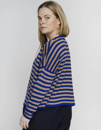A woman wearing a blue and beige striped knit top, shown from a side-back angle against a plain light gray studio background.
