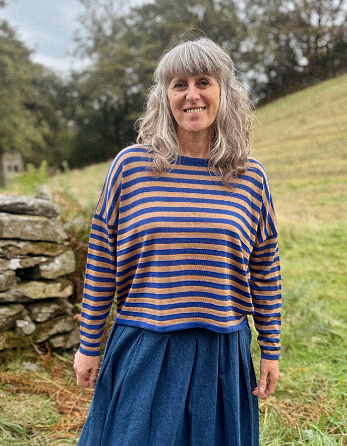 A smiling woman with gray hair stands outdoors wearing a blue and camel striped merino jumper with a denim pleated skirt, set against a grassy hill and stone wall background.