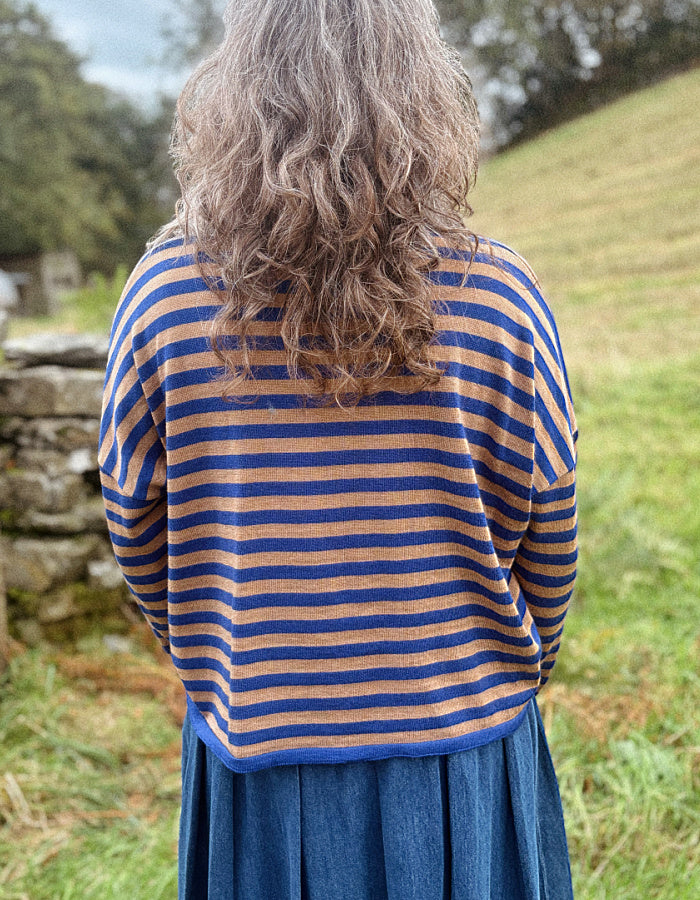 A person viewed from behind wearing a blue and camel striped knit merino top and a blue skirt, standing outdoors in a grassy field with a stone wall in the background.