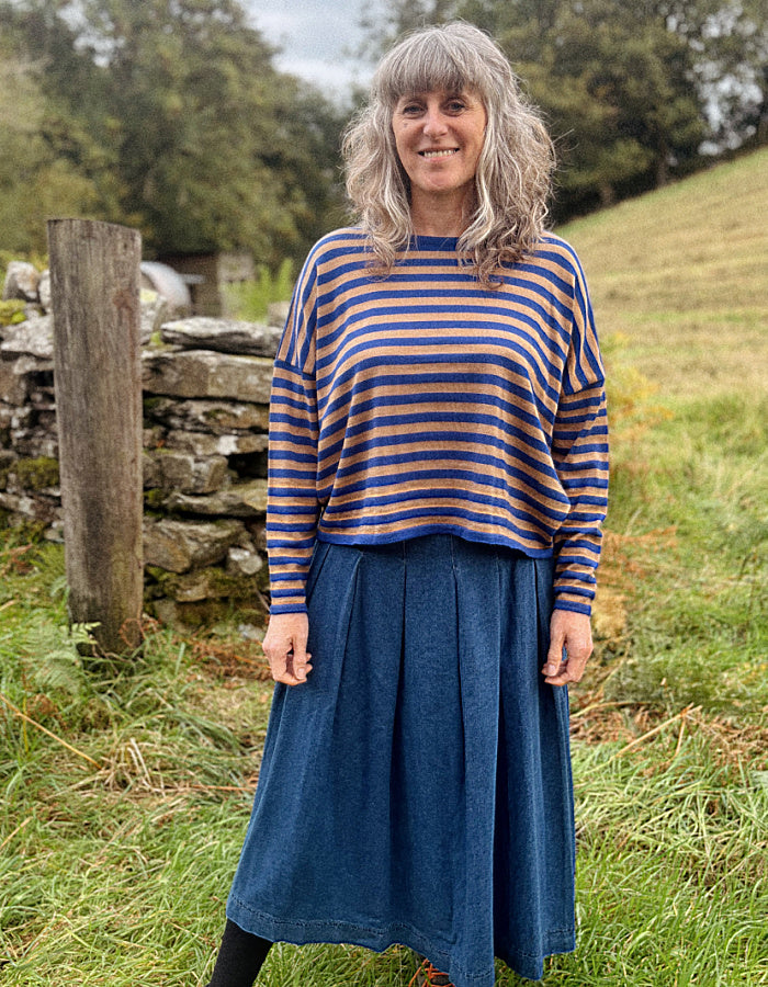 A woman standing outdoors in a grassy field wearing a blue and camel striped long-sleeve merino top and a blue denim skirt.