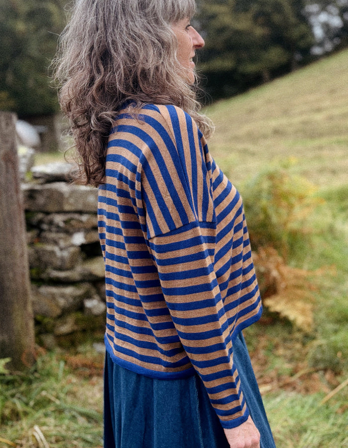A woman with long, wavy gray hair is shown from the side in an outdoor setting, wearing a blue and tan striped knit top and a dark blue skirt.