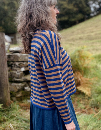 A woman with long, wavy gray hair is shown from the side in an outdoor setting, wearing a blue and tan striped knit top and a dark blue skirt.