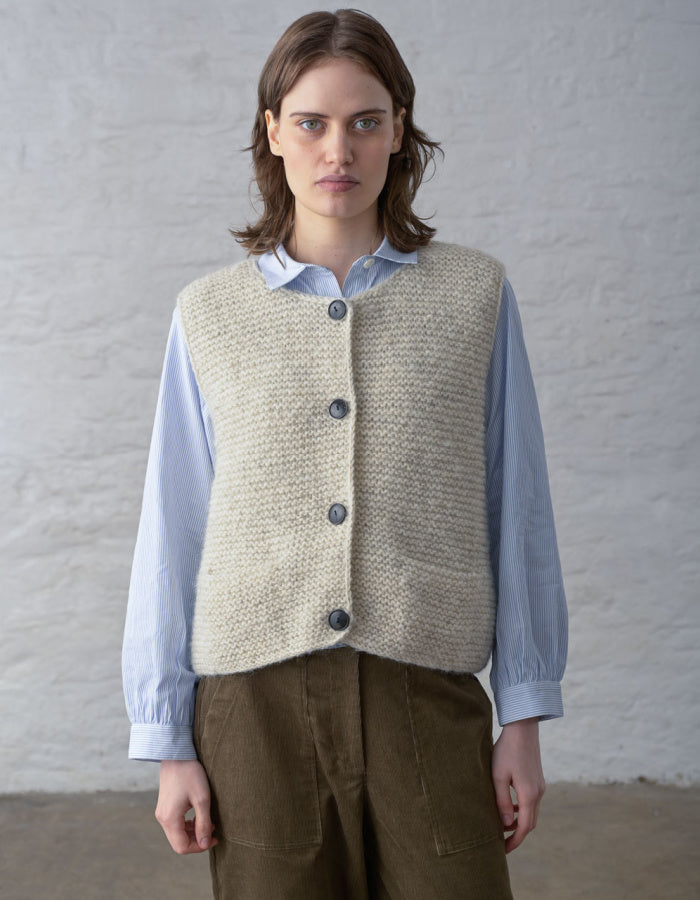 A model wearing a beige textured knit button-up vest over a light blue long-sleeve shirt and brown trousers, standing against a white brick wall.