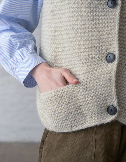 Close-up of a person wearing a chunky knit cream-colored vest with dark buttons over a light blue striped shirt and brown corduroy trousers.