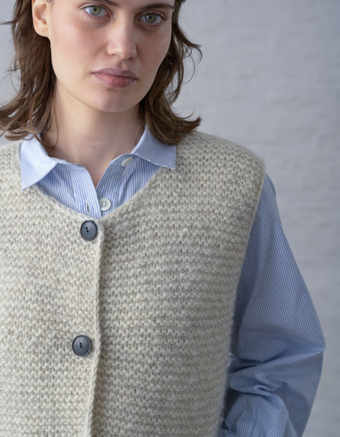 A close-up of a woman wearing a cream-colored knit vest over a light blue striped button-down shirt, set against a neutral gray background.