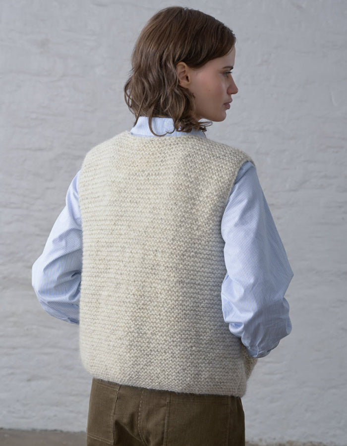 A woman shown from the back wearing a cream-colored textured knit sweater vest over a light blue long-sleeve shirt and brown trousers, standing against a white textured wall.