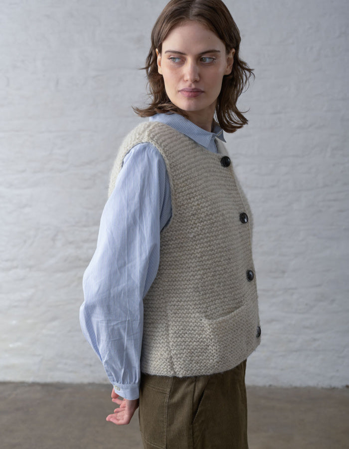 A woman wearing a beige textured knit vest over a light blue long-sleeve shirt and brown trousers, standing against a white brick wall.