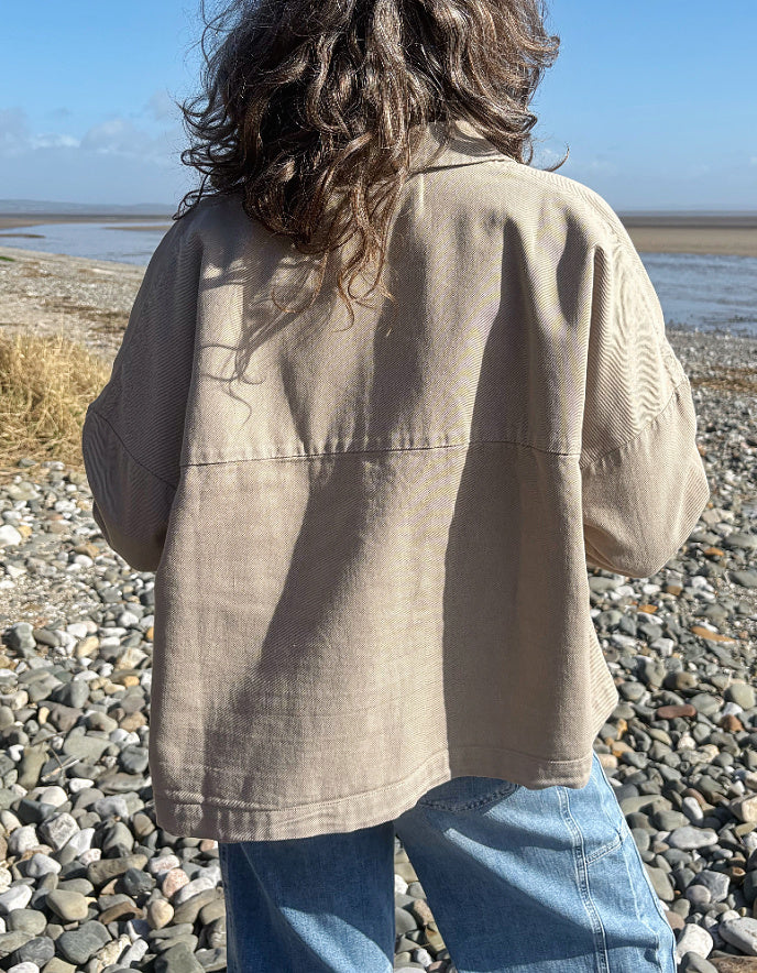 Person wearing a beige jacket standing on a pebbly beach with water and sky in the background