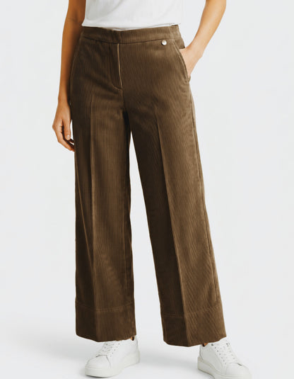 A woman wearing brown corduroy wide-leg cropped pants with a white top and white sneakers, shown from the waist down against a light grey studio background.
