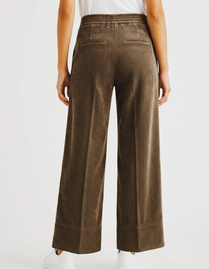A woman wearing brown corduroy wide-leg cropped pants with a white top and white sneakers, shown from the waist down against a light grey studio background.