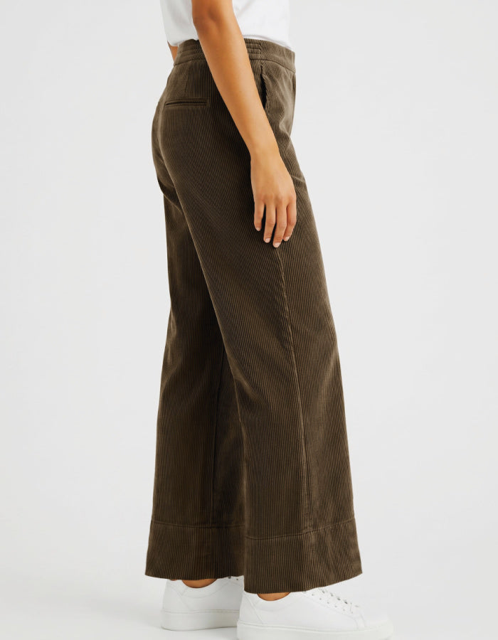 A woman wearing brown corduroy wide-leg cropped pants with a white top and white sneakers, shown from the waist down against a light grey studio background.