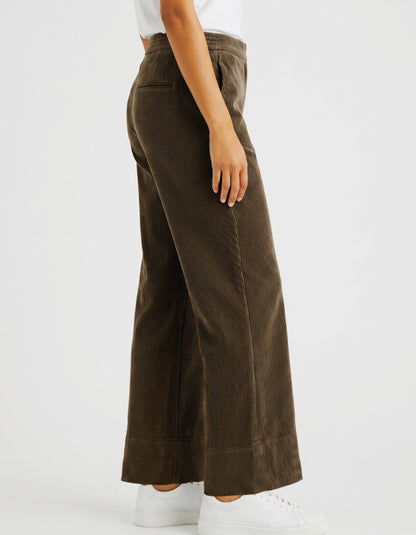 A woman wearing brown corduroy wide-leg cropped pants with a white top and white sneakers, shown from the waist down against a light grey studio background.