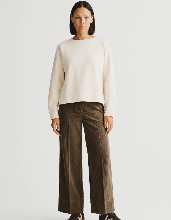 A model stands against a light gray studio background wearing a cream-colored long-sleeve knit sweater and dark brown wide-leg corduroy trousers.