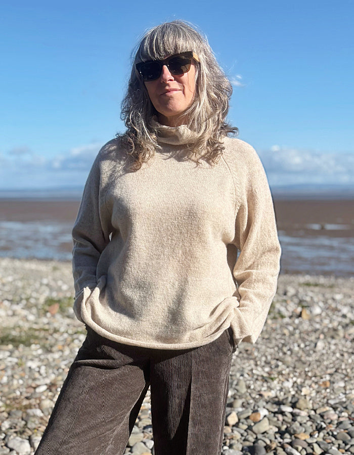 A woman with gray hair and sunglasses stands on a pebble beach wearing a cream-colored turtleneck sweater and brown corduroy trousers, with a blue sky and water in the background.