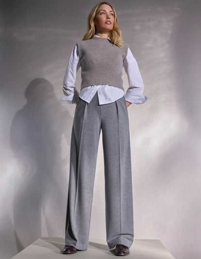 Woman wearing a gray sweater vest over a light blue shirt with grey wide-leg pants against a neutral background.