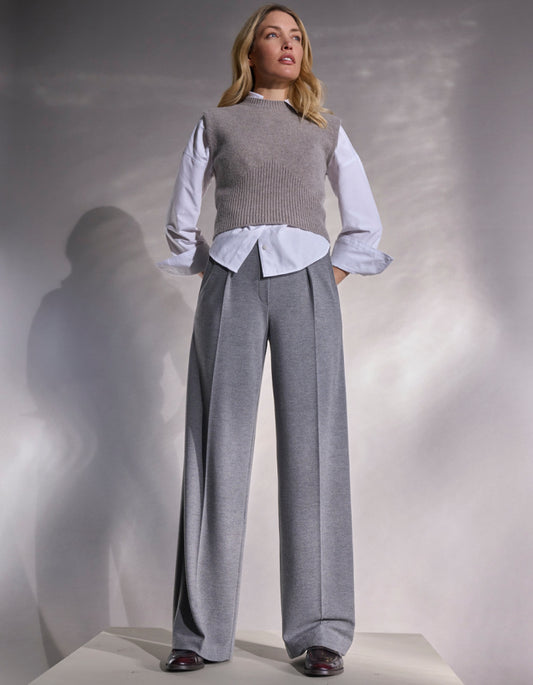 Woman wearing a gray sweater vest over a light blue shirt with grey wide-leg pants against a neutral background.
