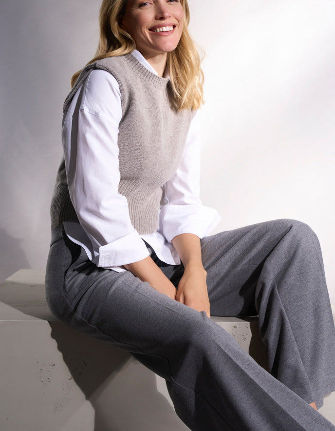 Woman wearing a gray sweater vest over a white shirt and gray pants, sitting on a white surface.