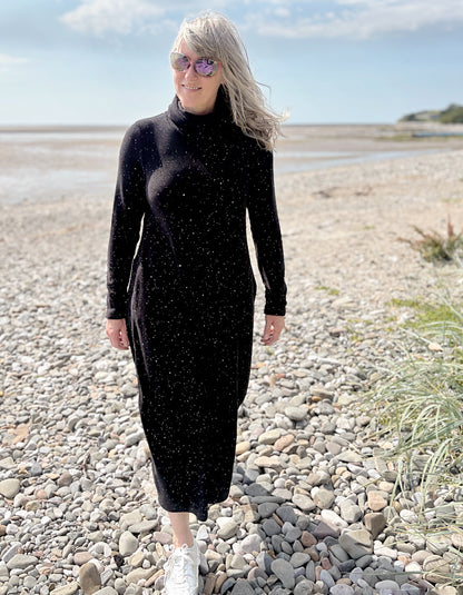 Person wearing a black dress with white speckled galactic print standing on a pebbly beach with a blue sky.