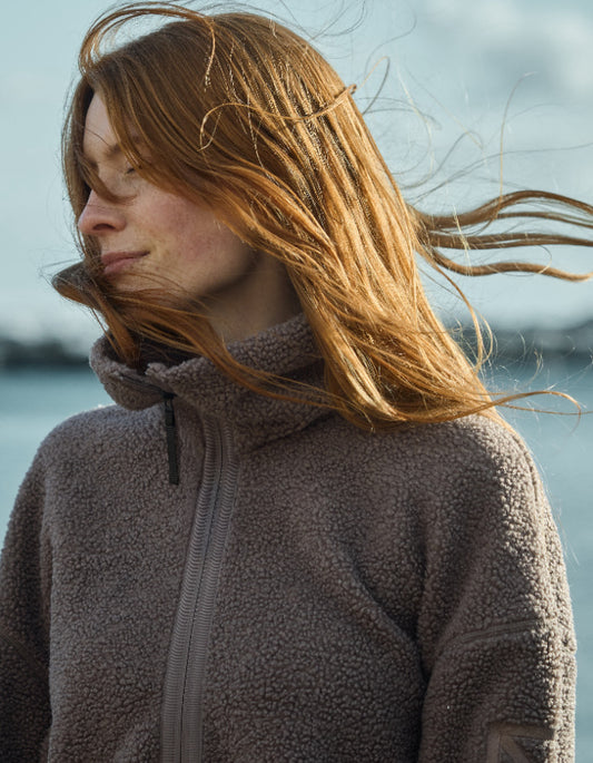 Woman with long red hair blowing in the wind wearing a beige brown full zip sherpa pile fleece jacket against a blurred natural background