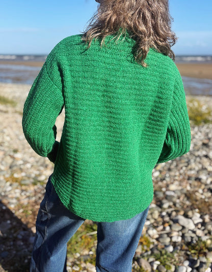 A person standing on a rocky beach wearing a bright green ribbed knit sweater and blue jeans, viewed from behind.