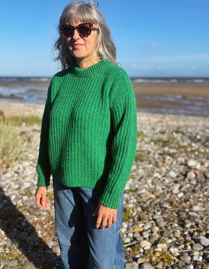 A woman with gray hair and sunglasses wearing a green ribbed knit sweater and blue jeans, standing on a pebble beach with water and sky in the background.