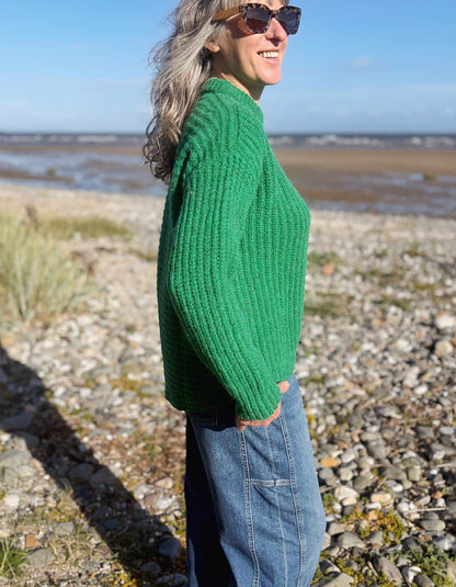 A woman wearing a chunky knit green sweater and blue jeans stands on a rocky beach shoreline, shown in profile against a blurred background of water and sky.