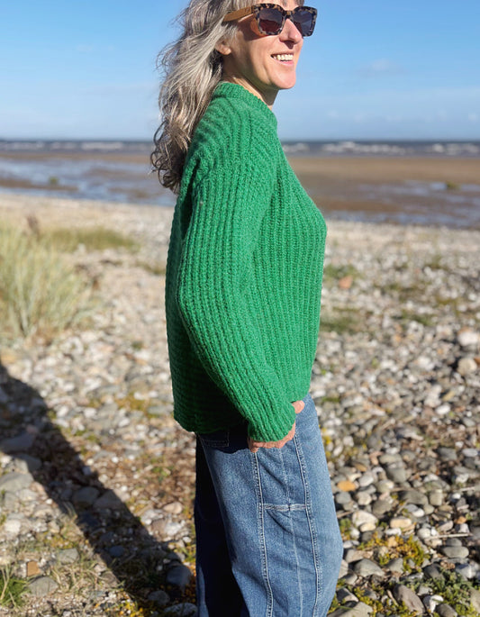 A woman wearing a chunky knit green sweater and blue jeans stands on a rocky beach shoreline, shown in profile against a blurred background of water and sky.