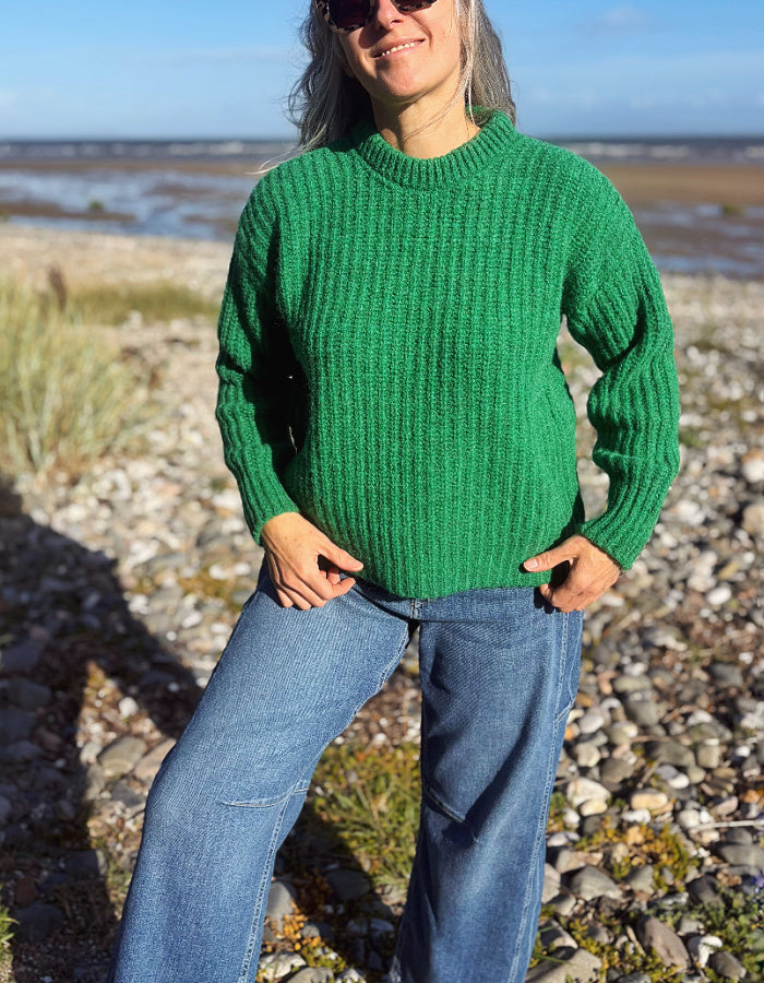 A woman wearing a vibrant green ribbed knit sweater and blue jeans stands on a rocky beach with the sea and sky in the background.