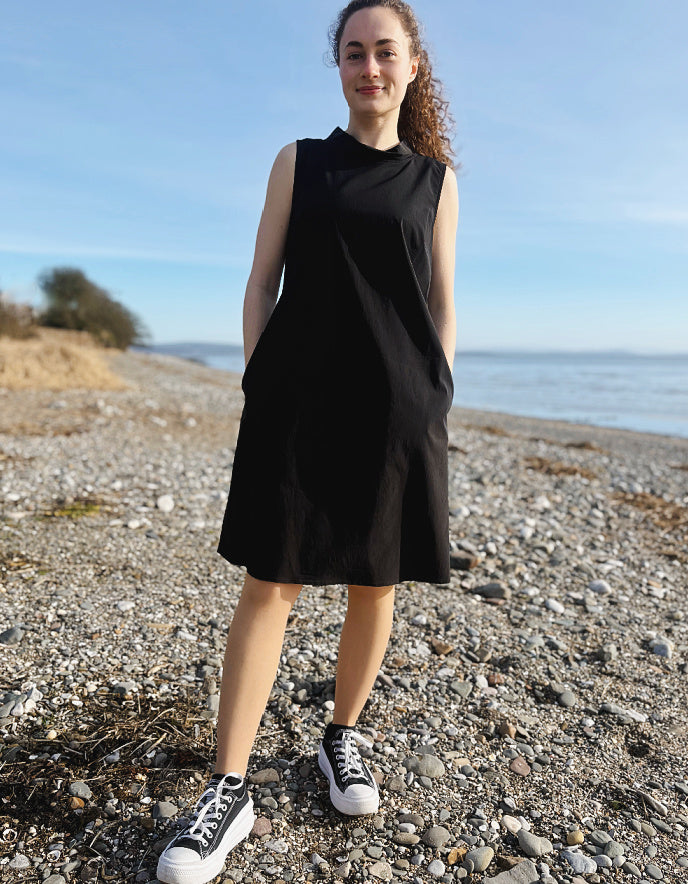 Woman wearing a black sleeveless dress standing on a pebbly beach with ocean and sky in the background.