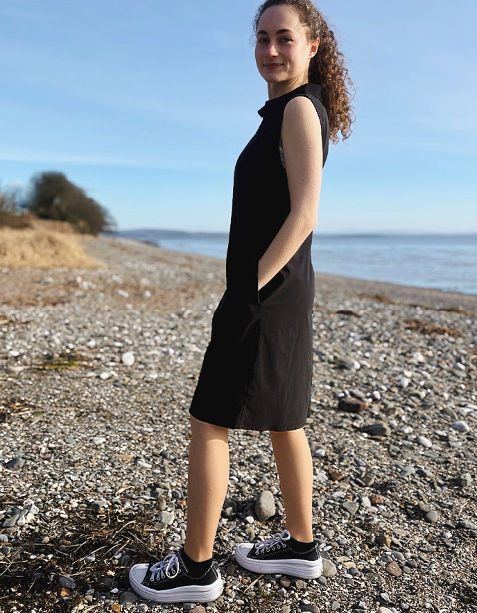 Person wearing a black sleeveless dress on a pebbly beach with ocean and sky in the background
