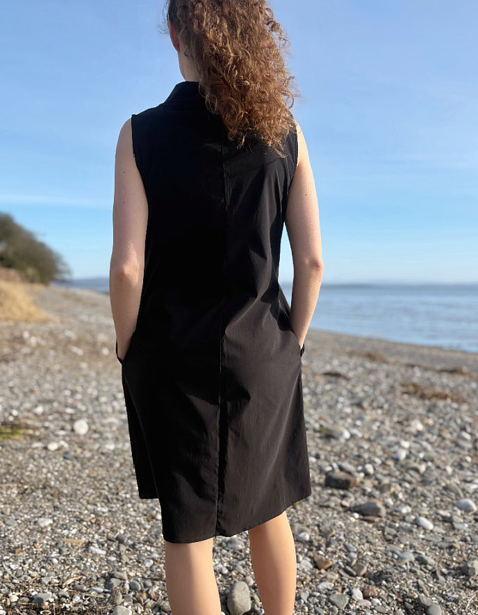 Person wearing a black sleeveless dress standing on a pebbly beach with ocean view.